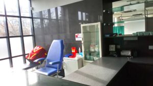 Wnętrze punktu szczepień. Niebieski fotel, oszklona szafka z medykamentami. Na leżance pod oknem czerwony plecak medyczny