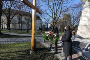 Poseł Urszula Rusecka oddaje hołd ofiarom Katynia oraz katastrofy Smoleńskiej. Asystent pani poseł kładzie biało-czerwoną wiązankę pod wysokim drewnianym krzyżem
