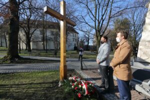 Poseł Rafał Bochenek i radny miejski Piotr Klimczyk. Spoglądają na drewniany krzyż stojący na wielickich Plantach. U stóp krzyża spoczywają biało-czerwone kwiaty