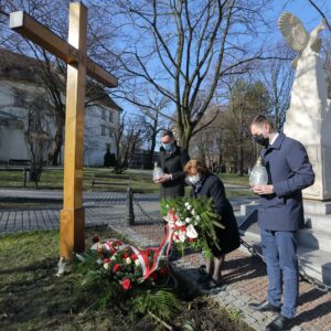 Przedstawiciele Zarządu Powiatu Wielickiego Aleksandra Ślusarek i Tomasz Broniowski oraz radny miejski Rafał Ślęczka. Pani Ślusarek składa u stóp krzyża wiązankę. Panowie trzymają zapalone białe znicze. Za ich plecami wielicki Pomnik Niepodległości zwieńczony orłem 