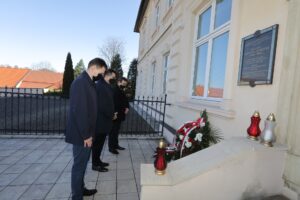 Tablica upamiętniająca Janusza Kurtykę na ścianie wielickiej siedziby IPN. Dyrektor Tomasz Broniowski oraz prezesi Paweł Nowak i Michał Roehlich składają hołd ofiarom katastrofy. Na niskim murku, poniżej tablicy, stoją zapalone znicze: dwa czerwone, jeden biały. Biało-czerwony wieniec oparto o otynkowaną na kremowo ścianę budynku, powyżej wieńca okno