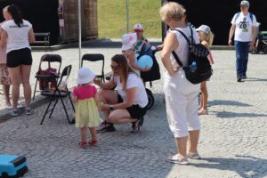 Gry i zabawy dla najmłodszych w tężni. Fizjoterapeutka kuca obok małej dziewczynki ubranej w różowo-żółtą sukienkę i biały kapelusik. Fizjoterapeutka pokazuje dziecku, jak zrobić mydlane bańki