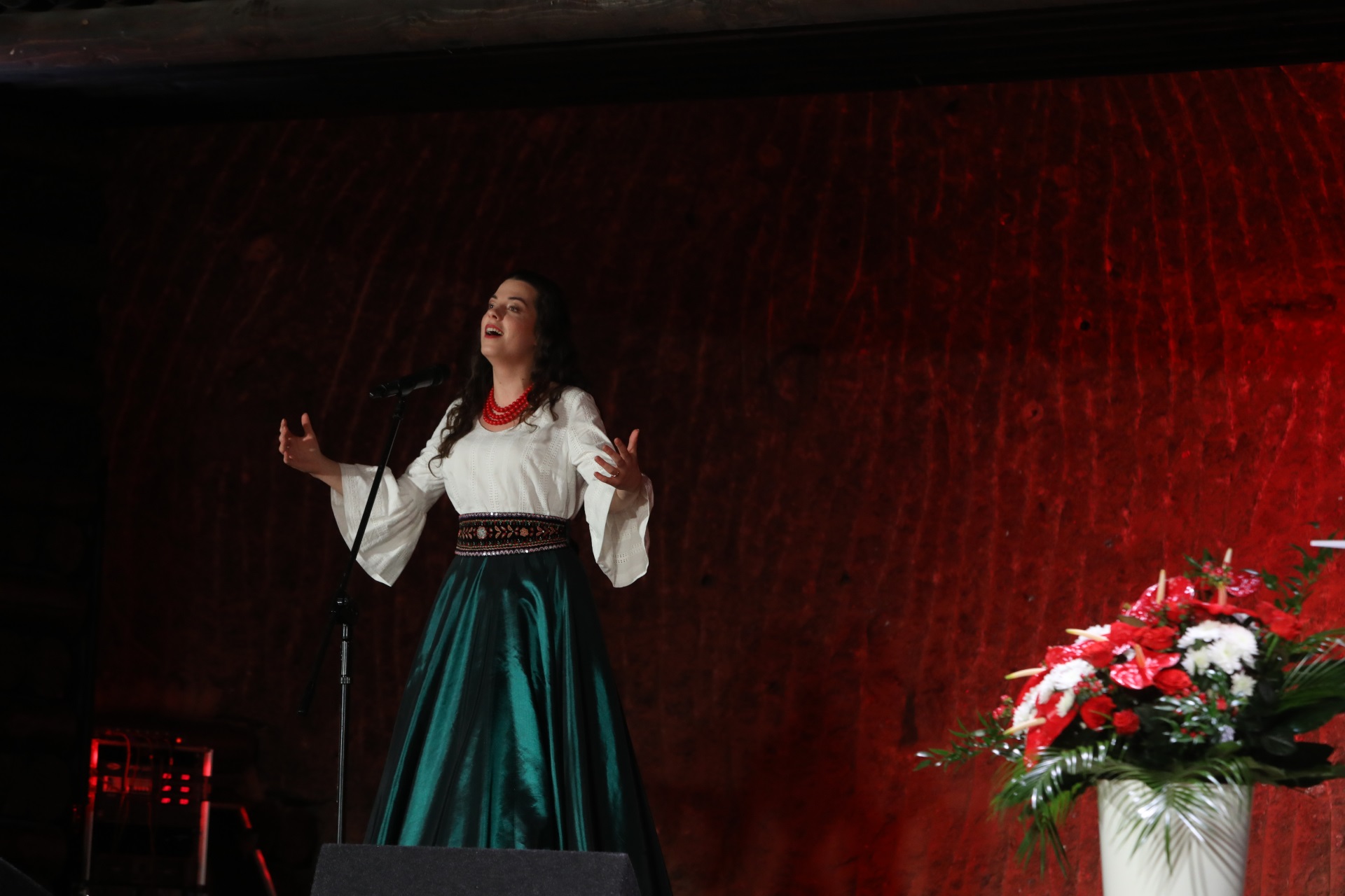 Sopranistka Wioletta Liber podczas solowego występu. Śpiewaczka jest ubrana w długą zieloną spódnicę, białą bluzkę. Ma na sobie ozdobny pas oraz czerwone korale