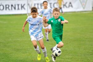 Zacięty pojedynek o piłkę. Na pierwszym planie zawodnik w biało-niebieskim stroju i zawodnik w stroju zielonym