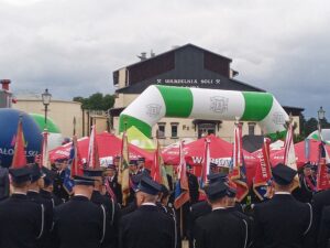 Strażacy a galowych mundurach. W tle poczty sztandarowe oraz budynek warzelni soli