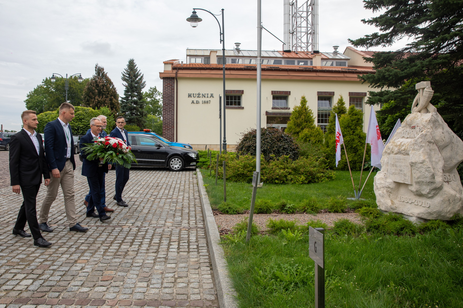 Delegacja składa kwiaty przy pomniku. W tle budynek z napisem kuźnia