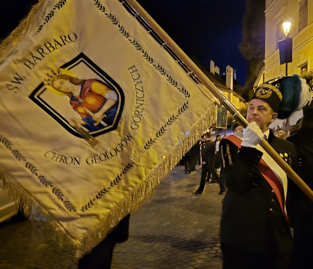 Haftowany sztandar z modlitwą do św. Barbary. Niesie go górnik w galowym mundurze. Zdjęcie zrobione po zmierzchu