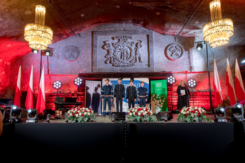 Górnicy na scenie śpiewają hymn. W tle rzeźbione w soli logo kopalni