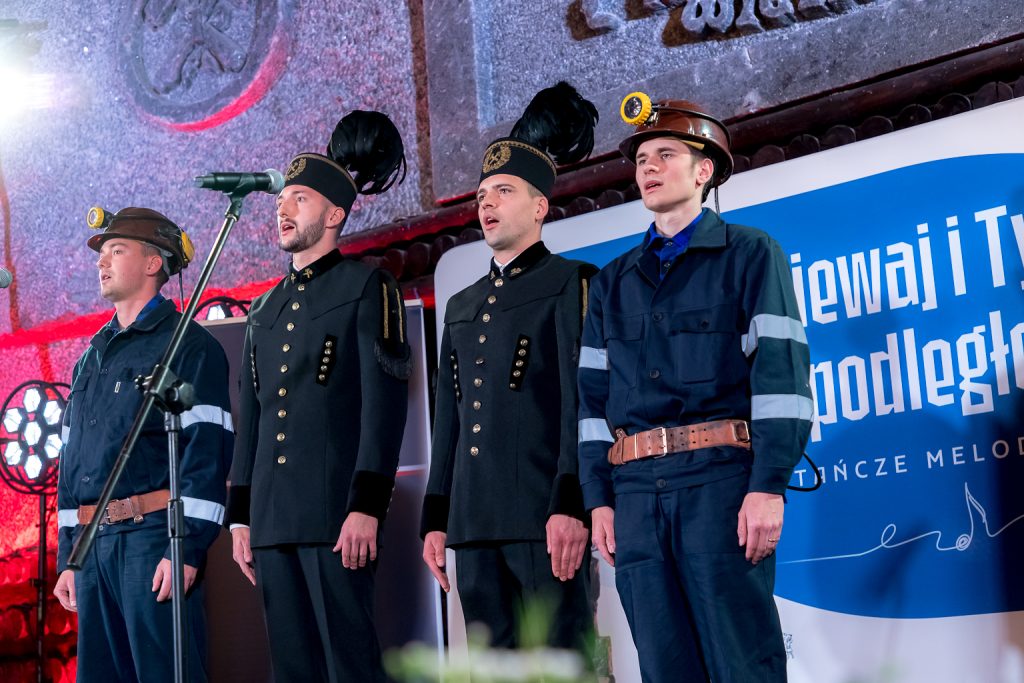 Górnicy na scenie. Śpiewają hymn. Dwóch w galowych mundurach, dwóch w codziennych górniczych strojach