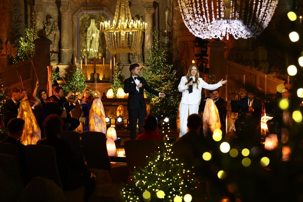 Dwójka wokalistów śpiewająca kolędę na tle ołtarza w kaplicy św. Kingi. Widoczny fragment żyrandola oraz ubrane świątecznie choinki