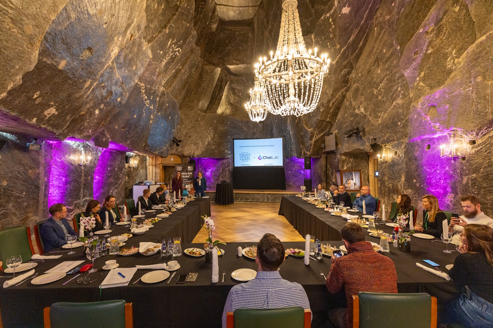 Widok na komorę Haluszka. Nakryte na czarno stoły ułożone w literę U. Nad głowami siedzących solny żyrandol