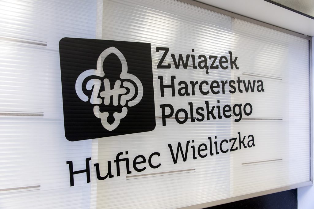 Czarny napis na białej ścianie: Związek Harcerstwa Polskiego Hufiec Wielicza. Napisowi towarzyszy znak lilii ze skrótem ZHP wewnątrz