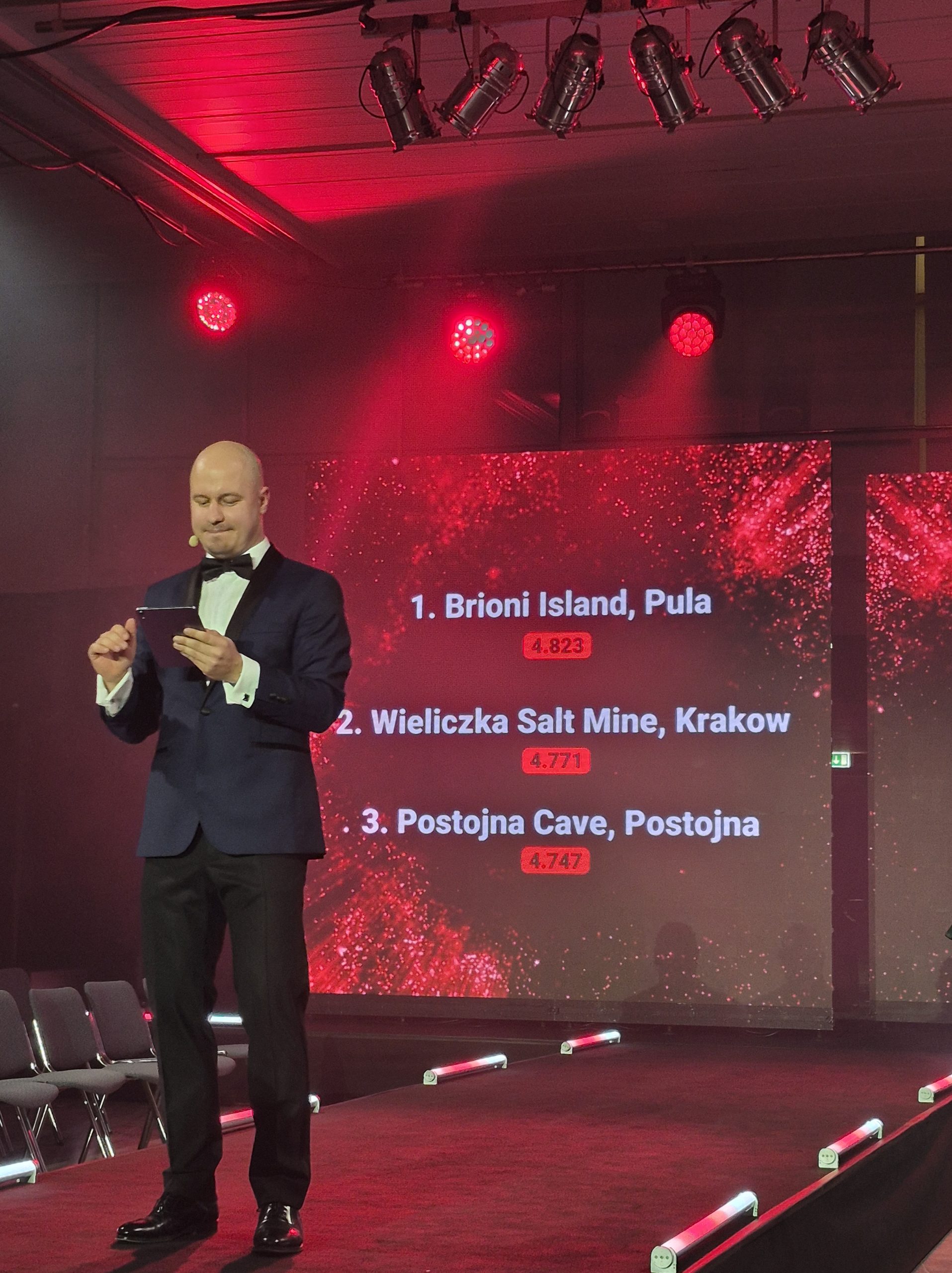 Prowadzący galę w galowym czarnym garniturze. Za jego plecami czerwono-czarny slajd z białymi napisami: z nazwami nagrodzonych miejsc i obiektów