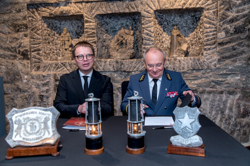 Prezesi podpisują umowę. Na stole dwie zapalone górnicze lampki oraz solne rzeźby: logo kopalni, logo Wisły Kraków