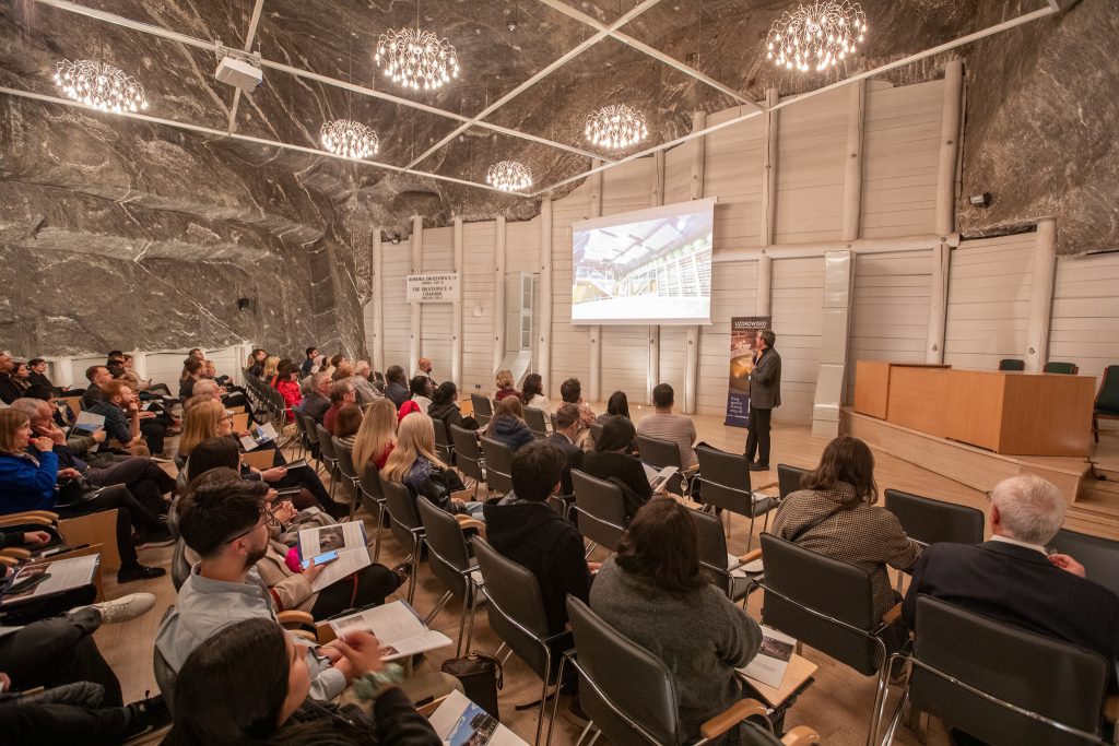 Podziemna sala konferencyjna oświetlona małymi żyrandolami. Uczestnicy spotkania słuchają wykładu