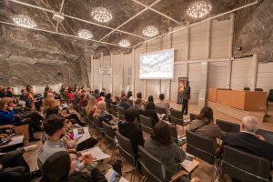 Podziemna sala konferencyjna oświetlona małymi żyrandolami. Uczestnicy spotkania słuchają wykładu