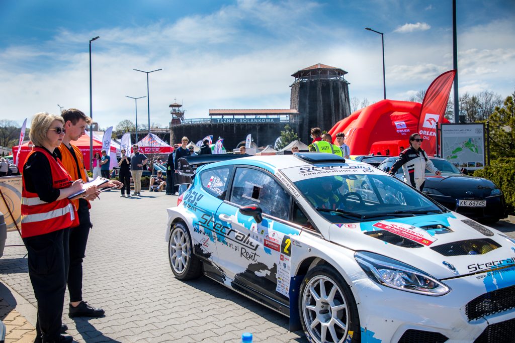 Rajdowy samochód - biało-niebieski, wyjeżdża z parkingu kopalni. W tle sylwetka tężni solankowej