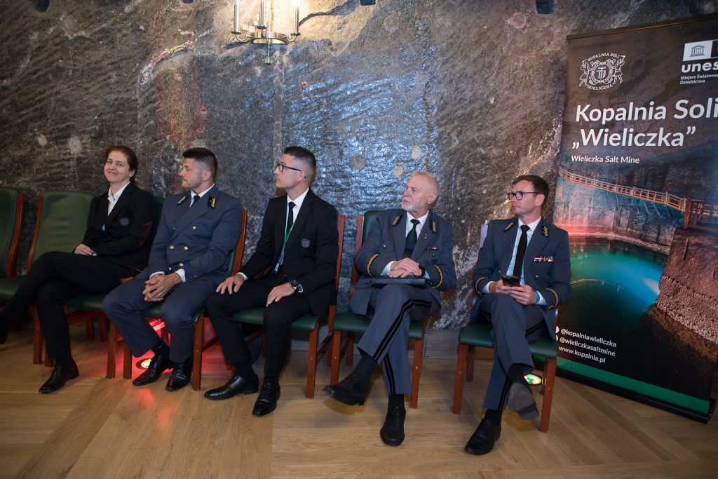 Przedstawiciele wielickiej kopalni. Czterech panów i jedna pani, siedzą na krzesłach ustawionych wzdłuż solnej ściany i słuchają wystąpień